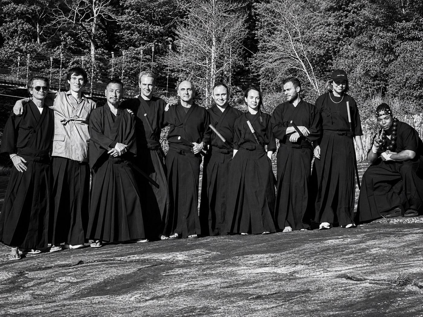 A training group in hakama with wooden swords, standing on a riverside rock in Japan.